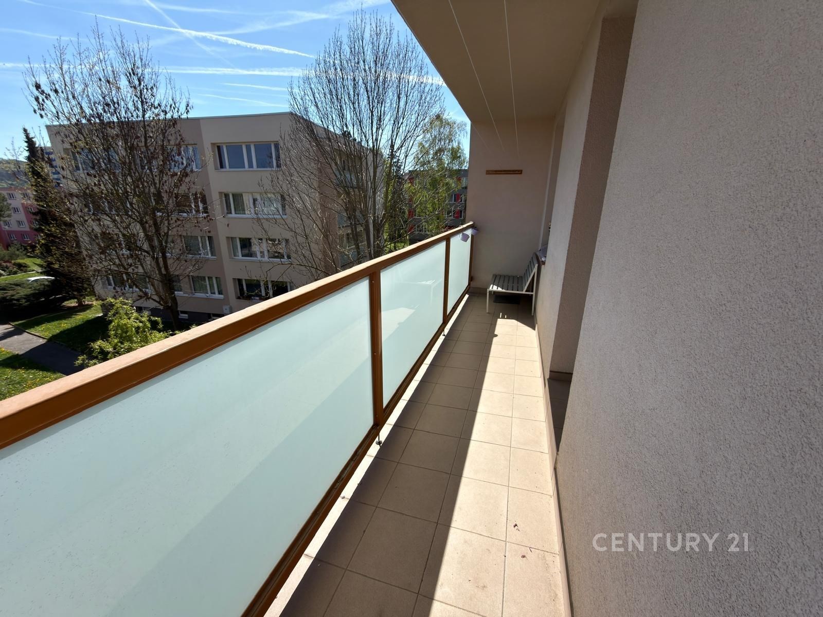 Prostorný balkon s dlažbou a moderním zábradlím nabízí výhled na klidné okolí a vzrostlé stromy, ideální pro relaxaci na čerstvém vzduchu.