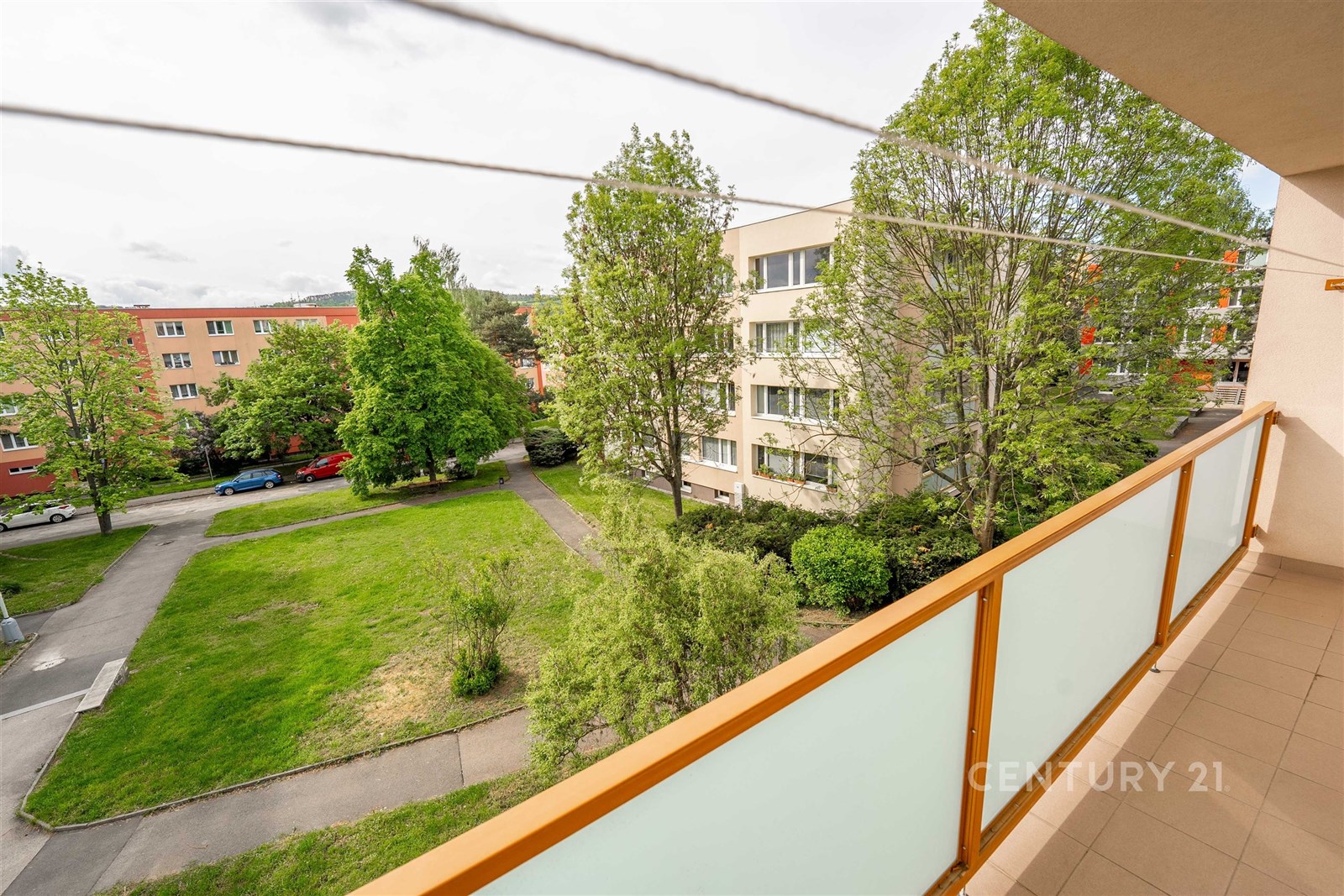 Balkon s moderním zábradlím nabízí výhled na klidnou čtvrť se zelení, nachází se u rekonstruovaného bytu v rezidenčním domě.