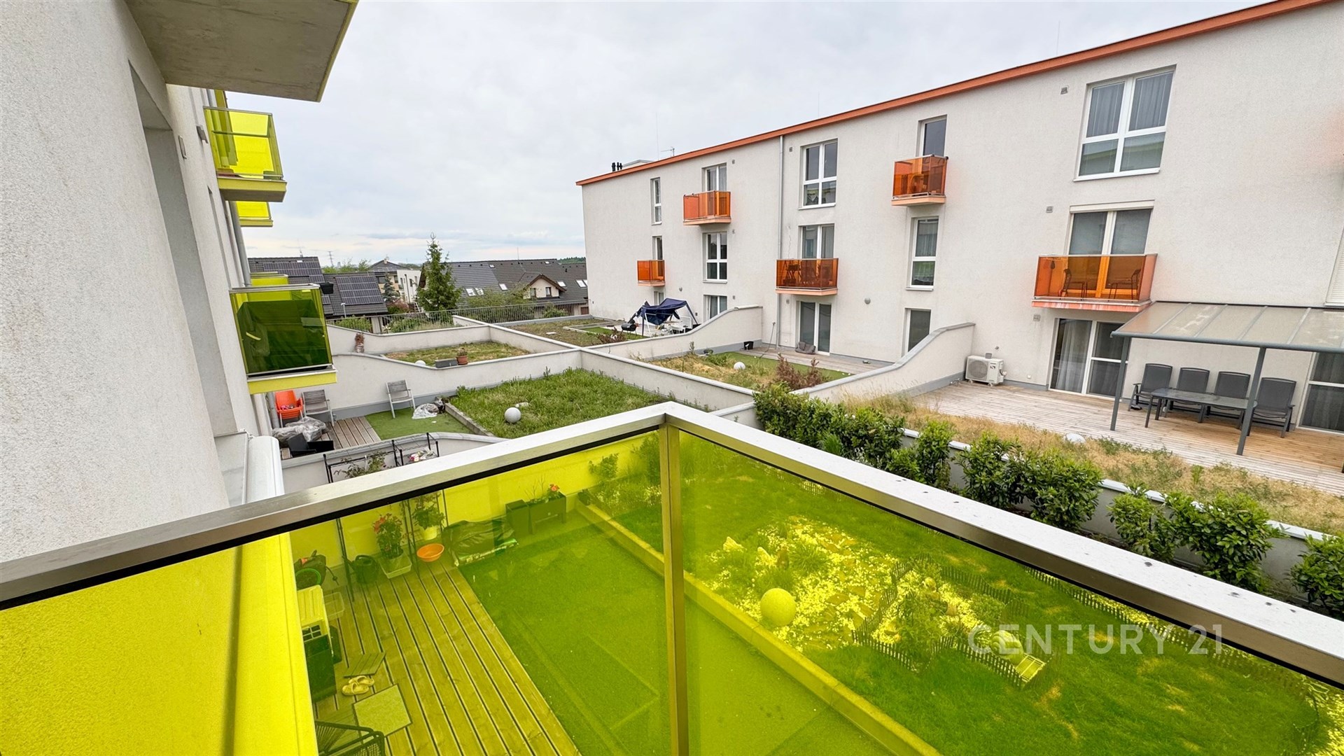 Balkon s moderním zábradlím, žluté sklo, výhled na udržovaný dvůr, novostavba bytového domu, klidná lokalita.
