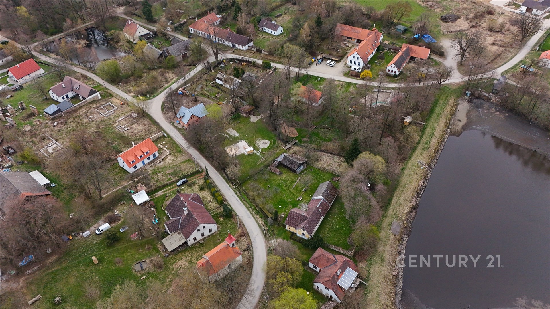 Letecký pohled na malebnou vesnici s tradičními domy, červenými střechami a přilehlým rybníkem, zasazenou do klidné krajiny.