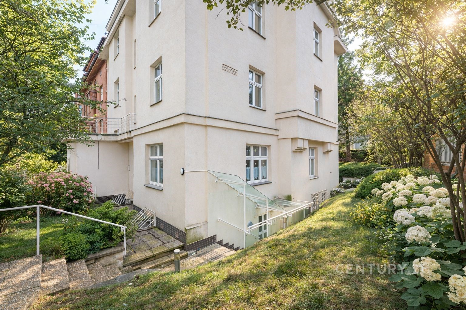 Exteriér zrekonstruovaného činžovního domu s upravenou zahradou, schodištěm a velkými okny, ideální pro klidné bydlení ve městě.