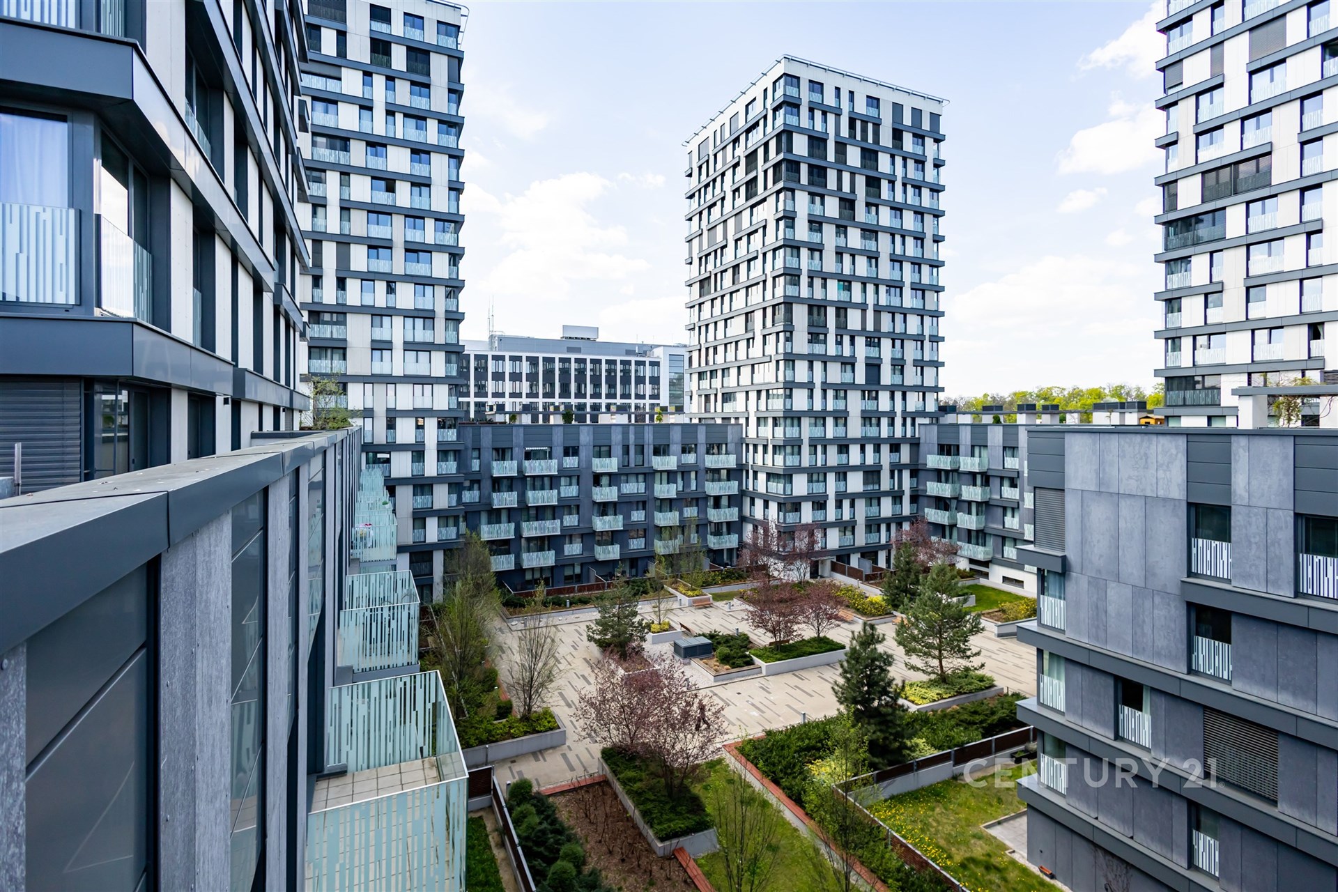 Výhled z balkonu na moderní bytový komplex s upraveným vnitroblokem, velkými okny a šedou fasádou.