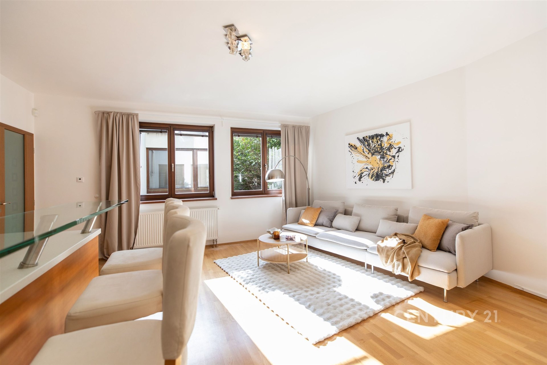 Světlý obývací pokoj s parketovou podlahou, velkými okny a moderním nábytkem, nabízí pohodlné posezení a elegantní designový styl.