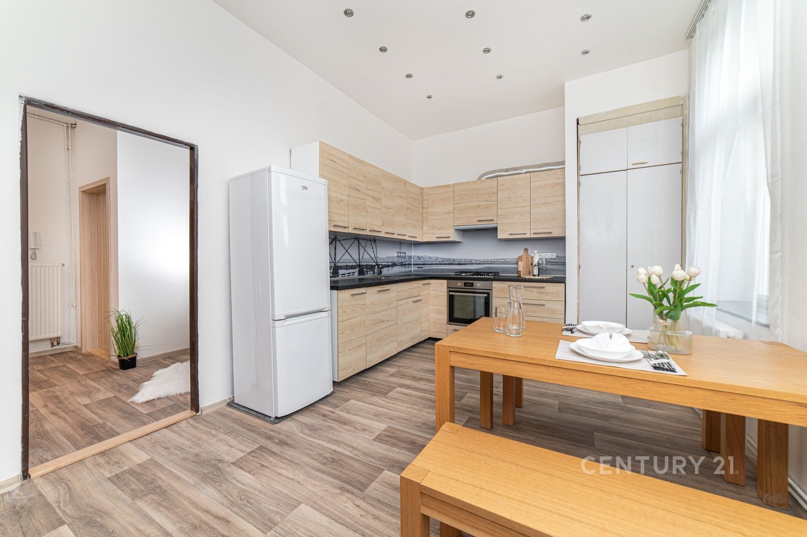 Moderní kuchyň s parketovou podlahou, světlými dřevěnými skříňkami a vestavěnými spotřebiči, vedle ní dřevěný jídelní stůl s květinami.