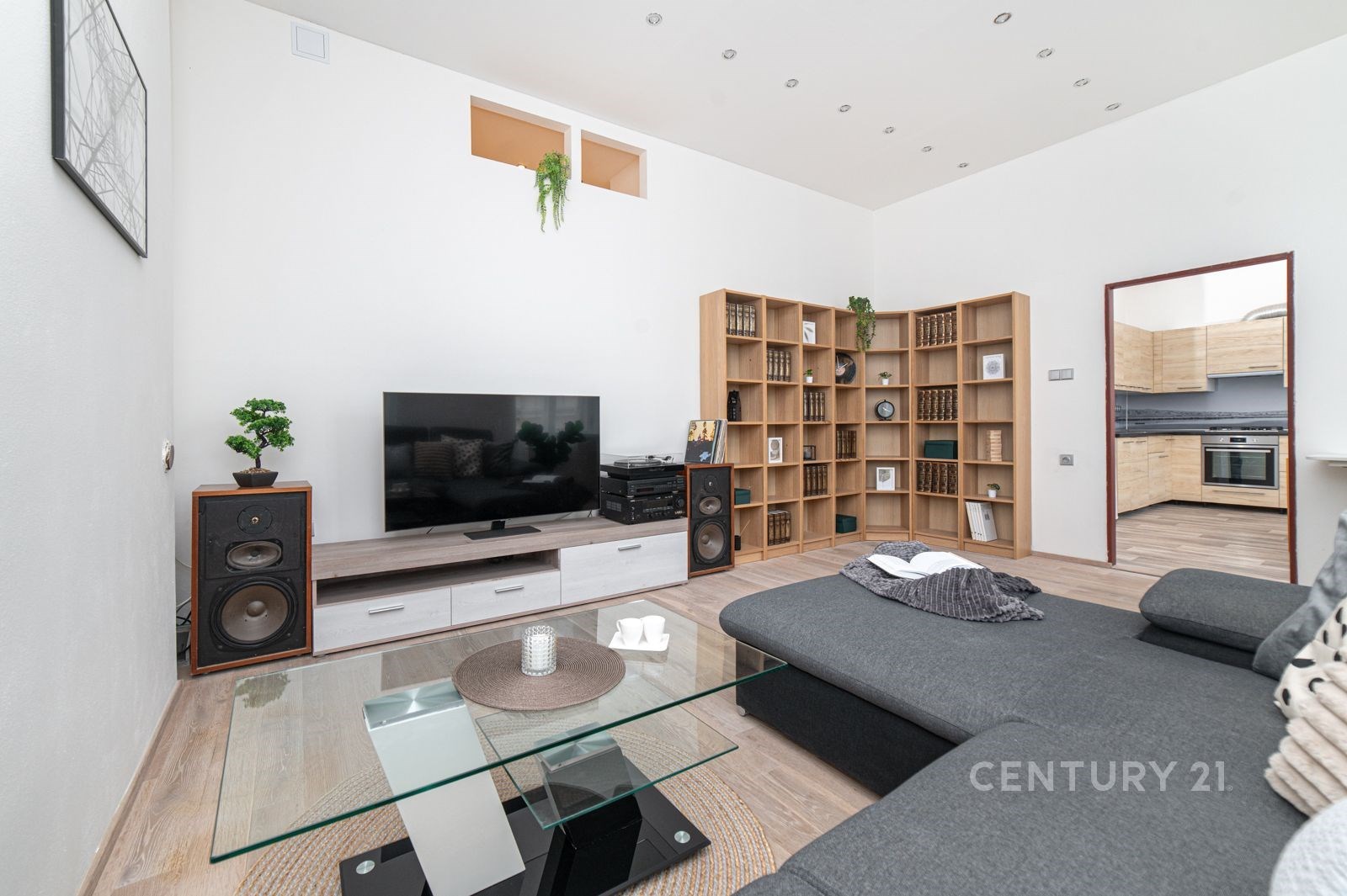 Světlý obývací pokoj s parketovou podlahou, moderním nábytkem a velkou knihovnou, propojený s kuchyní v pozadí, ideální pro rodinné bydlení.