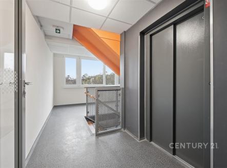 Chodba s moderním výtahem, kovovým zábradlím a velkými okny, která zajišťují dostatek denního světla, na podlaze je praktická dlažba.