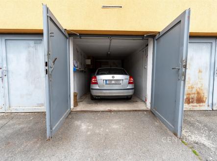 Prázdná garáž s kovovými dveřmi a betonovou podlahou, v pozadí zaparkované auto, vhodná pro parkování nebo skladování
