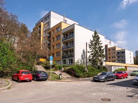 Fasáda bytového domu s moderními prvky, rekonstruovaná s novými balkonovými zábradlími, obklopena zelení a parkovacími místy.