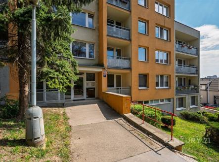 Vchod do bytového domu se širokou přístupovou cestou, moderní fasádou a balkóny. Nachází se v klidné lokalitě s upravenou zelení.