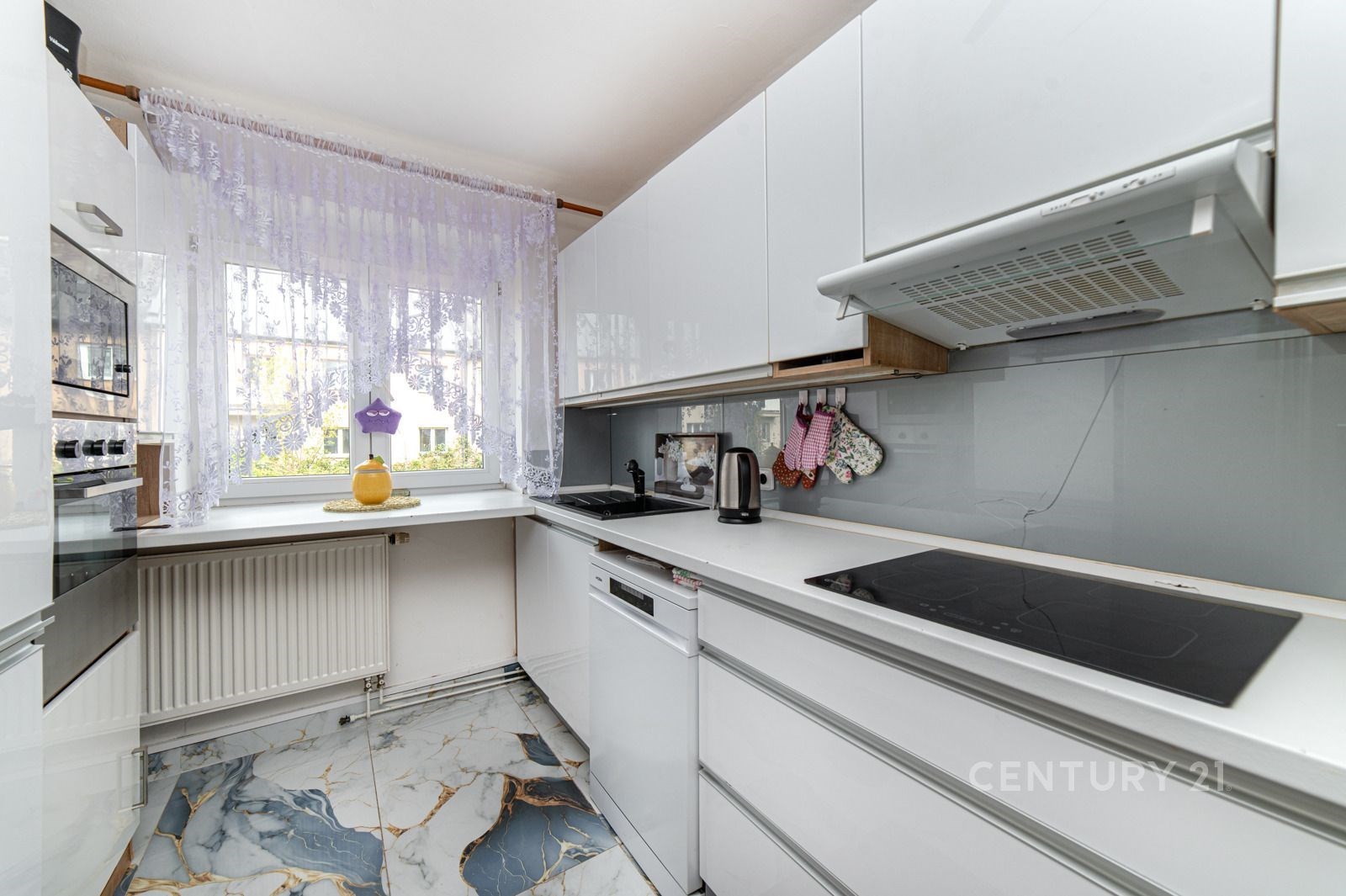 Moderní kuchyň s lesklými bílými skříňkami, elegantní mramorovou dlažbou a velkými okny zajišťujícími dostatek světla.