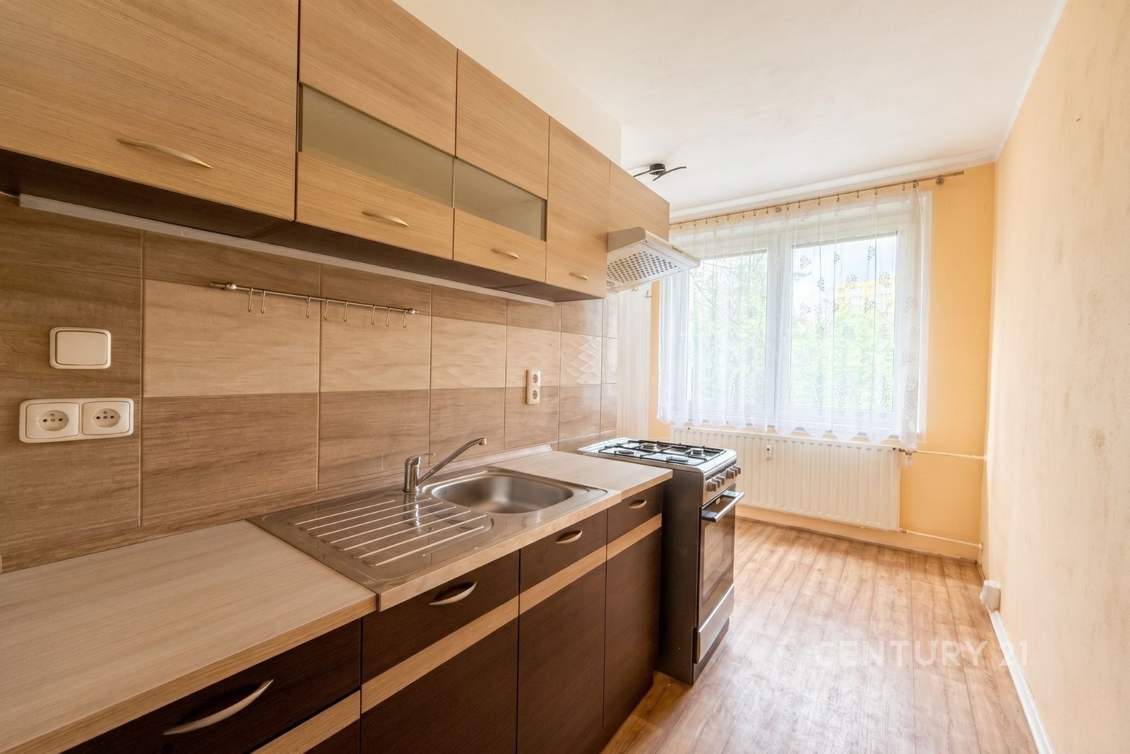 Kompaktní kuchyň s dřevěnou skříní, moderním vybavením a parketovou podlahou, velké okno přináší přirozené světlo.
