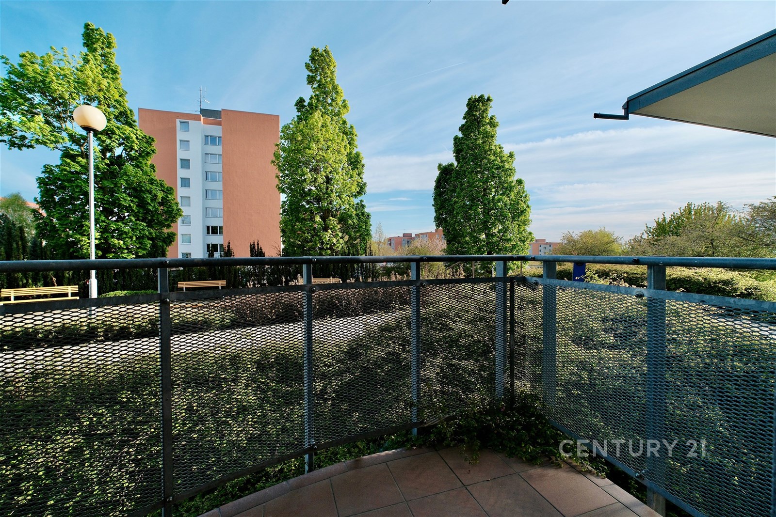 Balkon s keramickou dlažbou a moderním zábradlím nabízí výhled na zeleň a okolní bytovou zástavbu, ideální pro relaxaci v klidném prostředí.