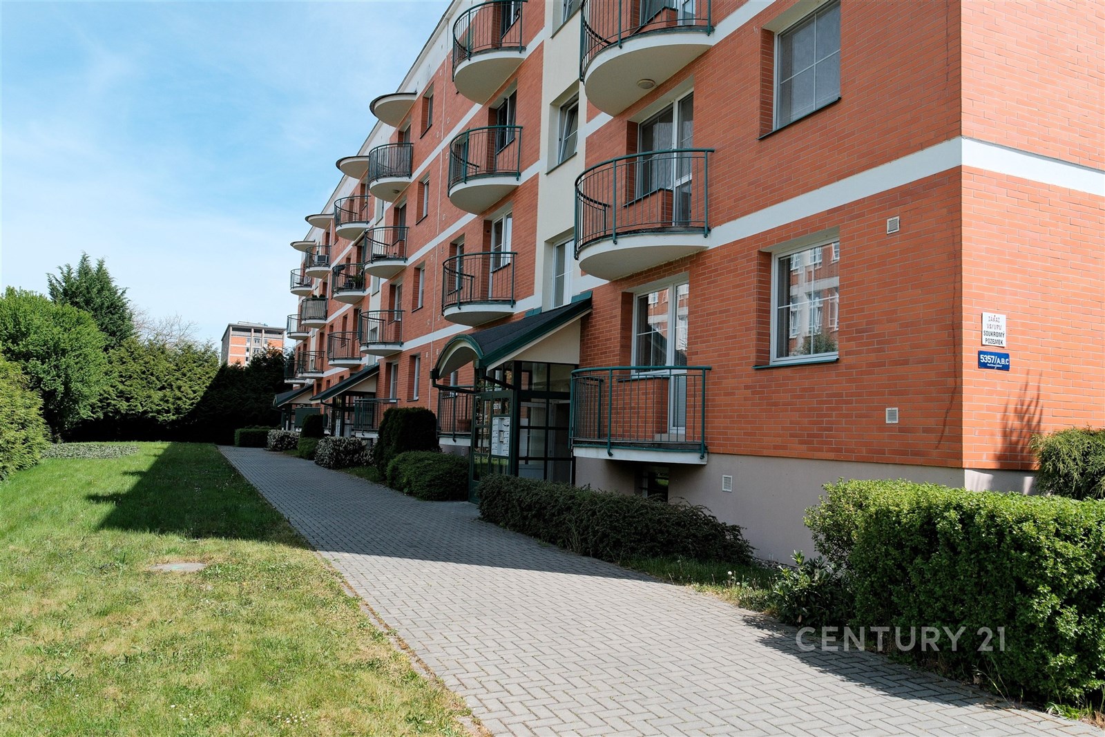 Červená cihlová fasáda moderního bytového domu s balkonem a zámkovou dlažbou, udržovaný trávník a zahrada, klidná lokalita.