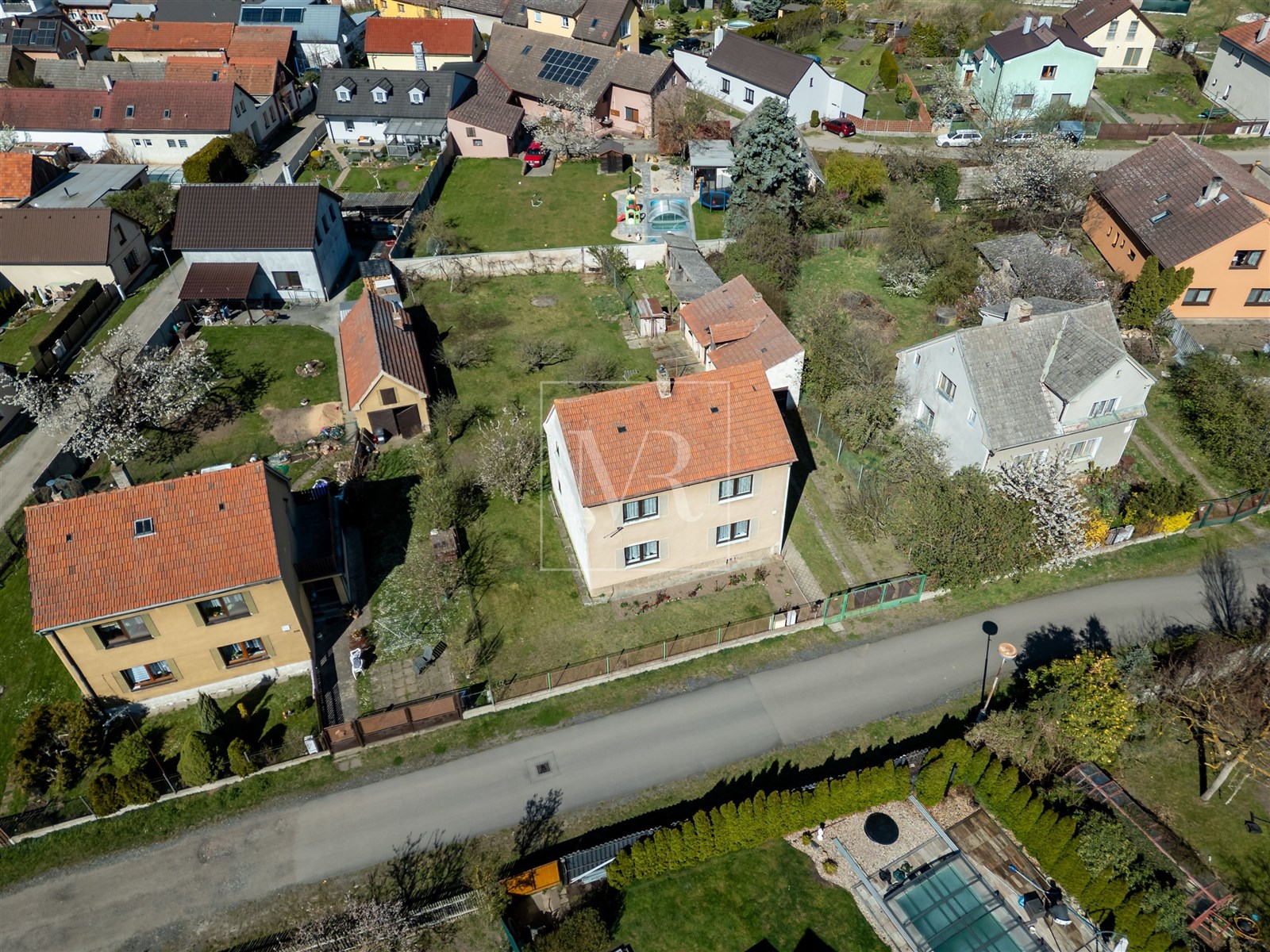 Letecký pohled na rezidenční čtvrť se zahradami a rodinnými domy, každý s tradiční střechou a zelení kolem.