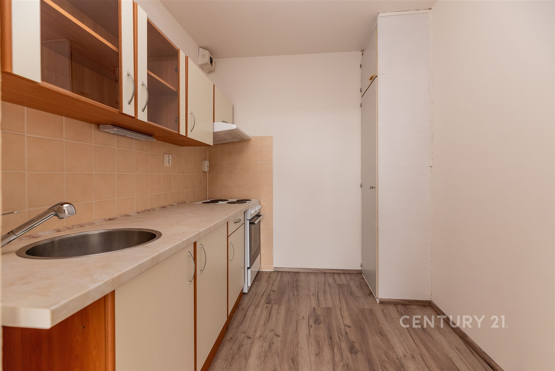 Úzká kuchyň s parketovou podlahou, dřevěnými skříňkami a světlou pracovní deskou, moderní keramická dlažba na zdi kolem sporáku.