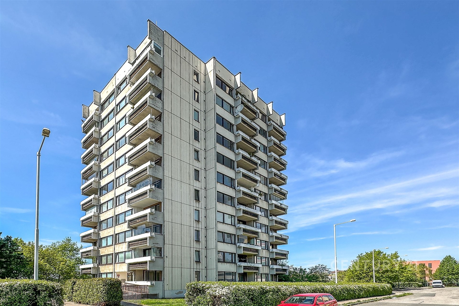 Vysoký bytový dům s balkony z roku 2000,  udržovaná zelená plocha, světlý exteriér, klidná lokalita, výhled na okolí, příjemné prostředí.