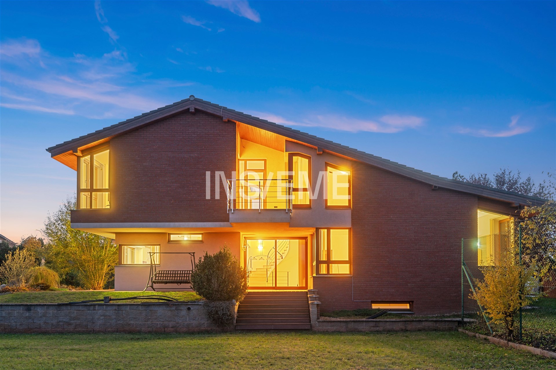 Dvoupodlažní rodinný dům s cihlovou fasádou a velkými okny, moderní design, upravená zahrada, večerní osvětlení vytváří útulnou atmosféru.