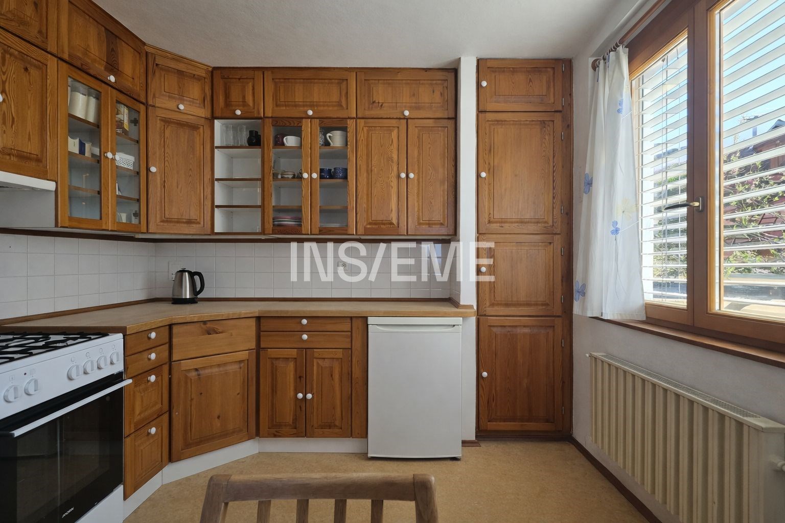 Kuchyň s dřevěnými skříňkami, velkými okny a bílými dlaždicemi, světlý prostor s moderním vybavením a vinylovou podlahou.