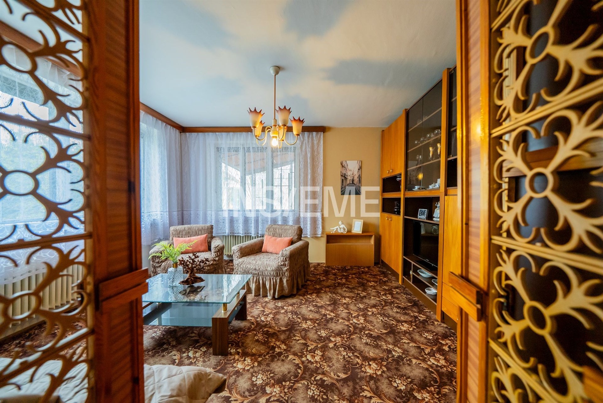 Světlý obývací pokoj se starožitným nábytkem, velká okna, dřevěné detaily a koberec s vzorem, zachovalý retro styl.