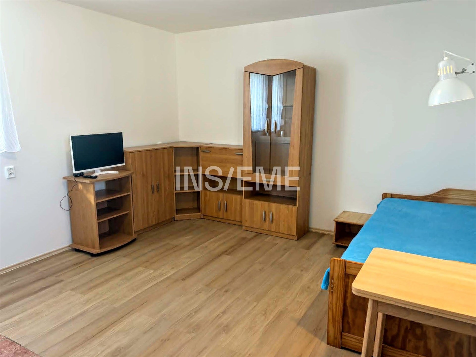 Světlý pokoj s dřevěnou podlahou, vestavěnou skříní a moderním nábytkem, včetně pracovního stolu a televize.