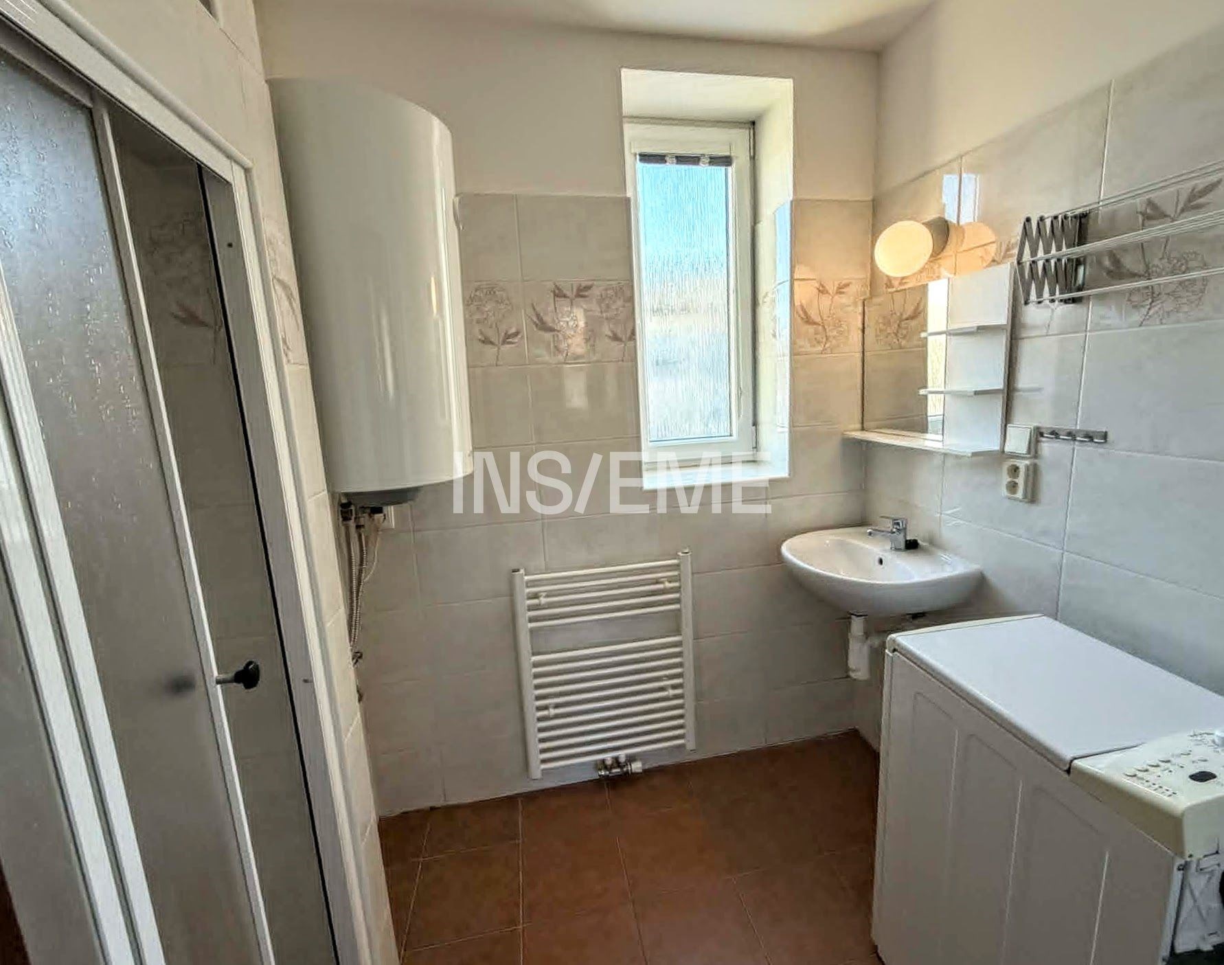 Koupelna s dlažbou a bílými stěnami, vybavená umyvadlem a moderním bojlerem, světlá díky velkému oknu.