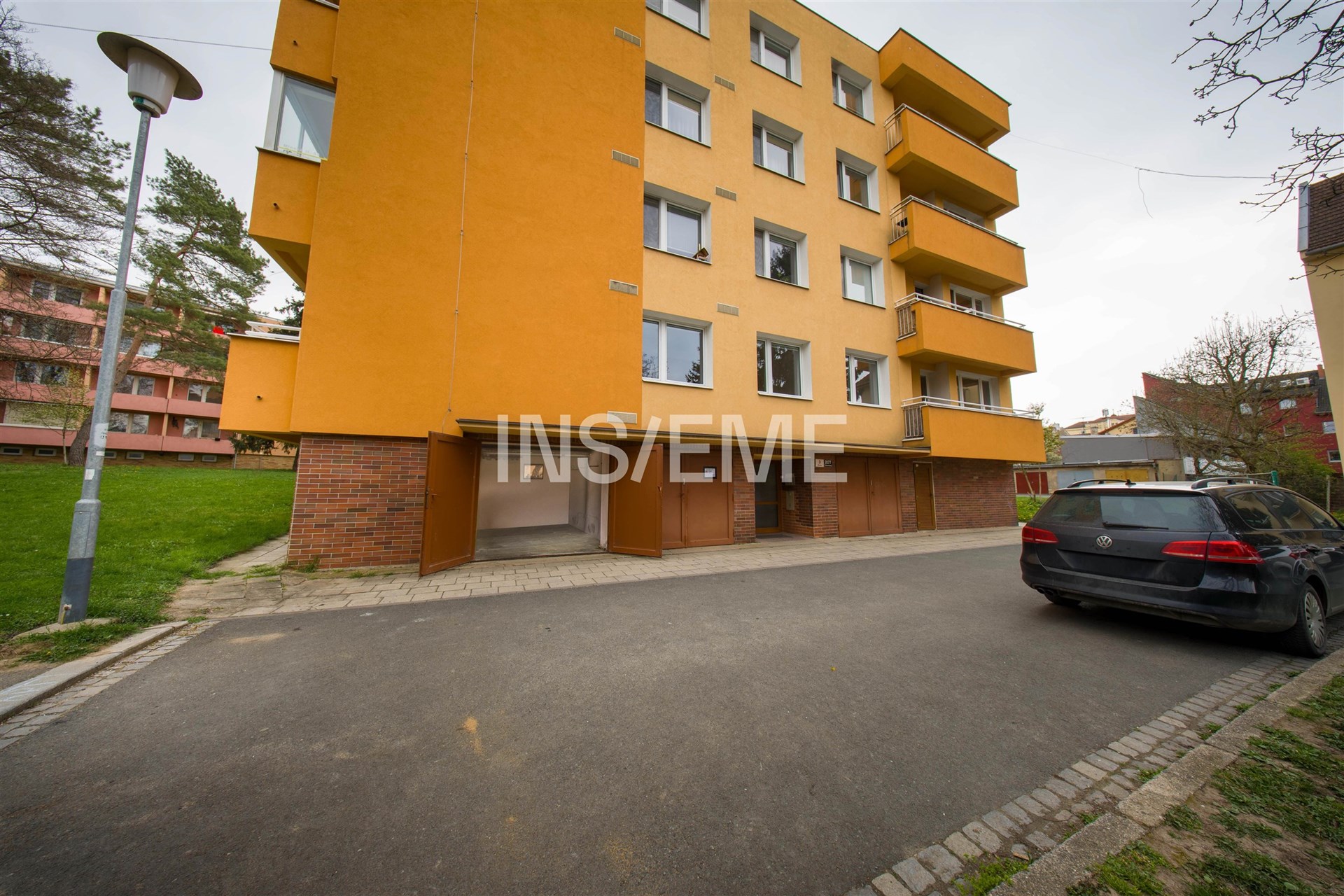 Vchod do bytového domu s moderní fasádou a nezastřešeným parkováním, obklopený zelení a přilehlou asfaltovou cestou pro parkování.