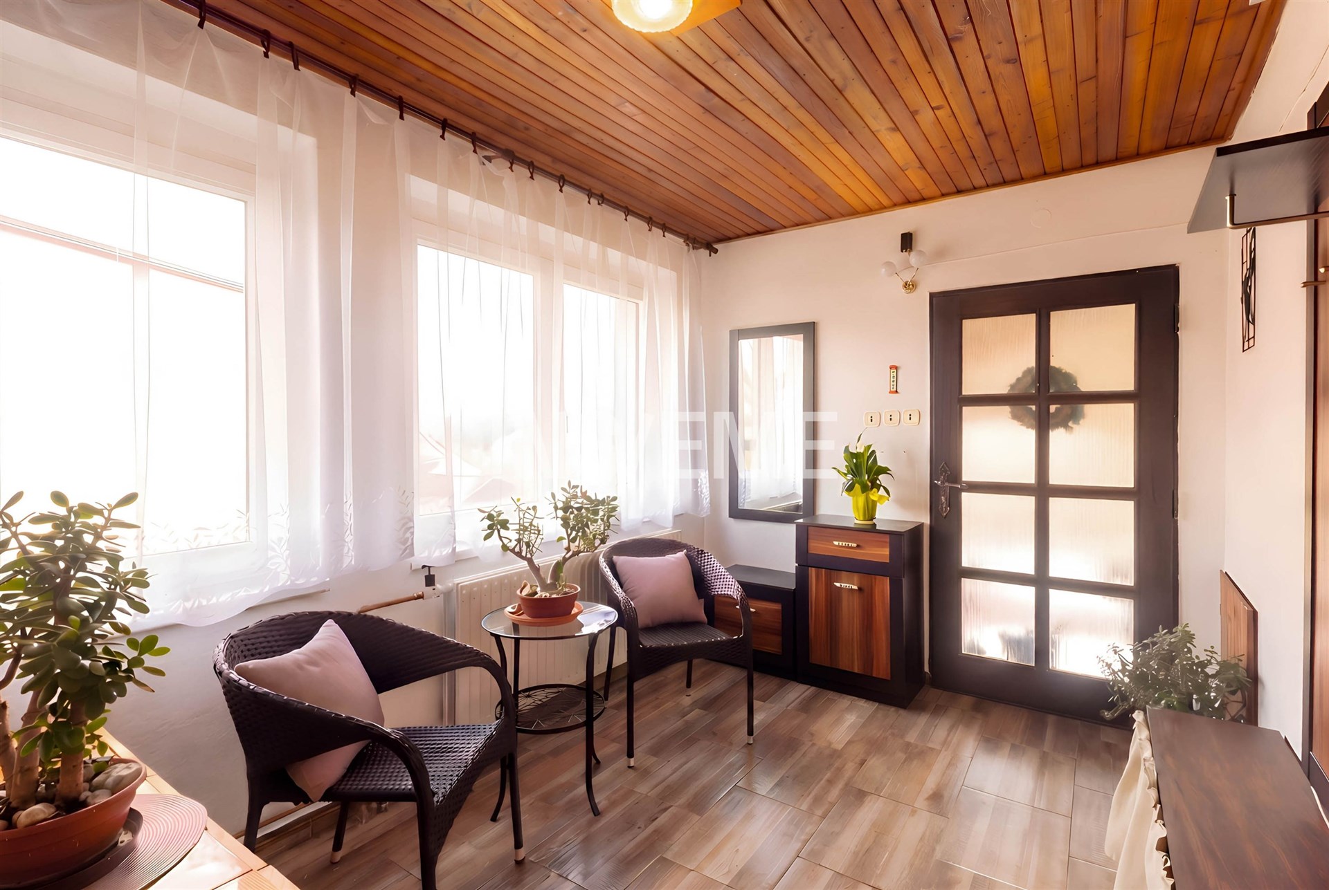 Světlá veranda s dřevěným stropem, dlažbou a velkými okny, moderní design s ratanovým nábytkem a rostlinami.