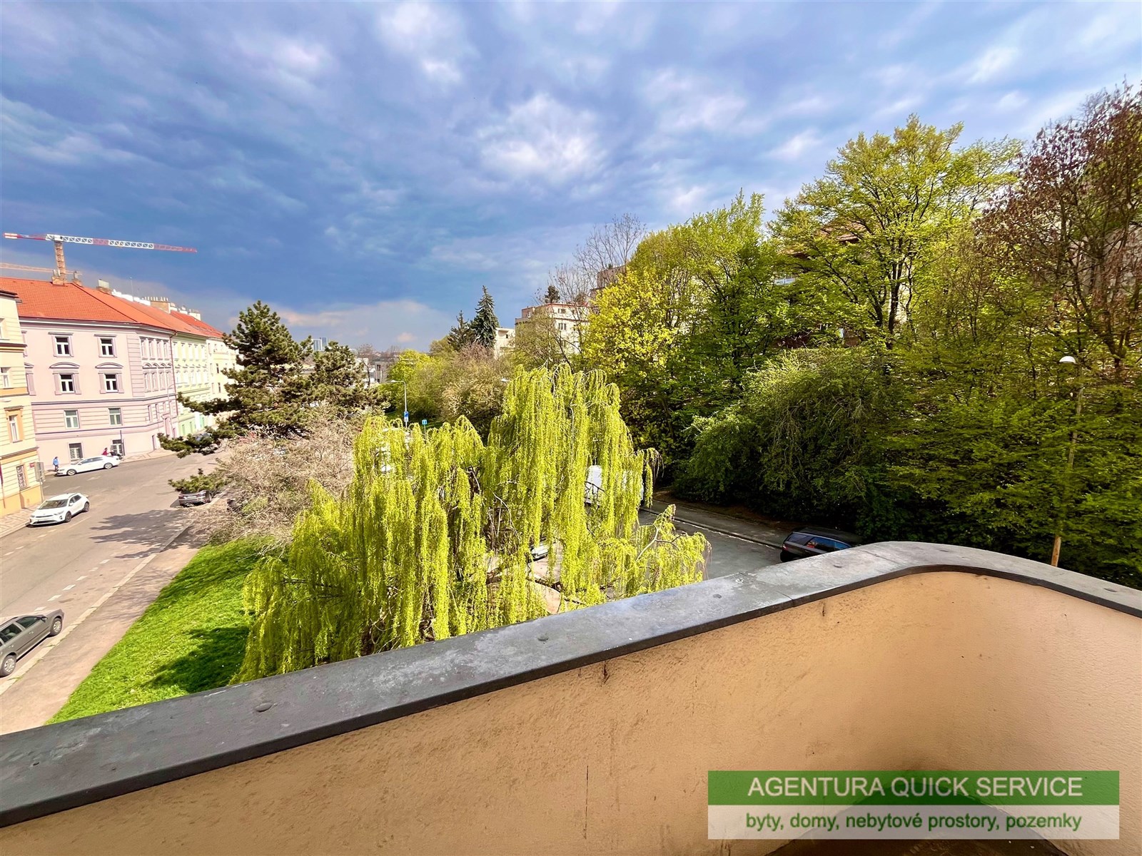 Pohled z balkonu s kovovým zábradlím nabízí výhled na klidnou zelenou lokalitu s množstvím stromů a parků, ideální pro relaxaci.