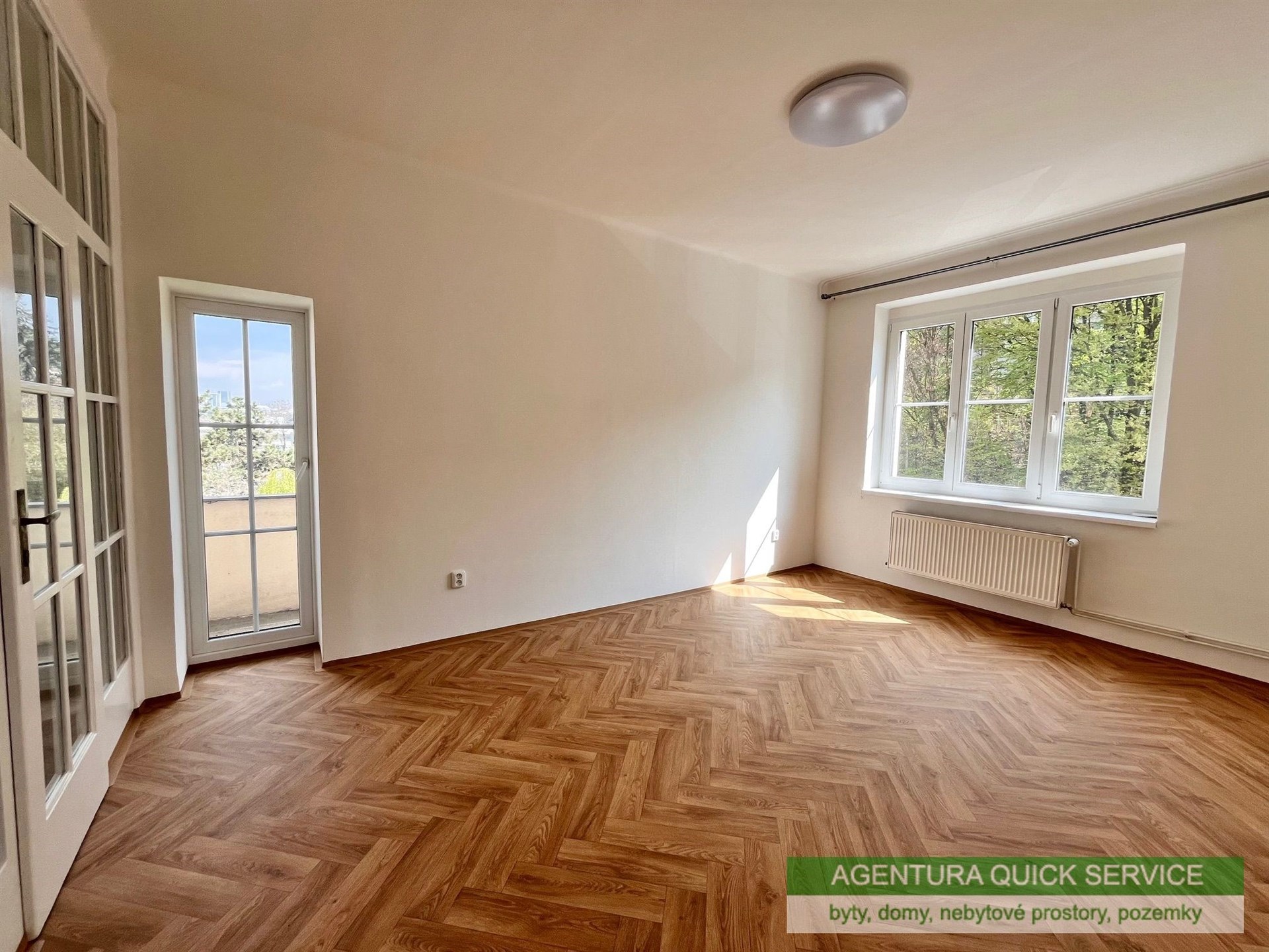 Světlý obývací pokoj s parketovou podlahou, velká okna poskytují výhled do zahrady, moderní interiér s prosklenými dveřmi na balkón