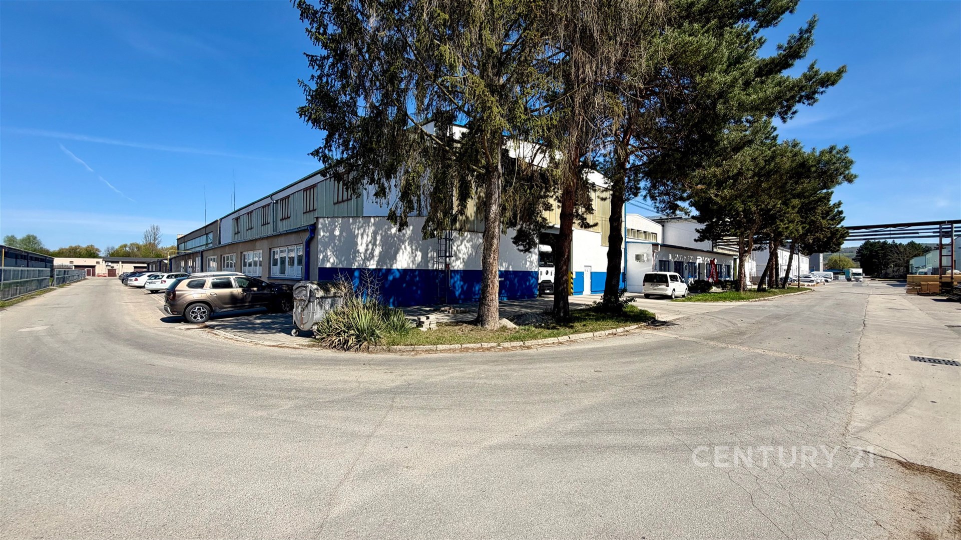Průmyslový areál s budovou v pozadí, obklopený parkujícími automobily, asfaltová cesta, vzrostlé stromy, oplocený pozemek.