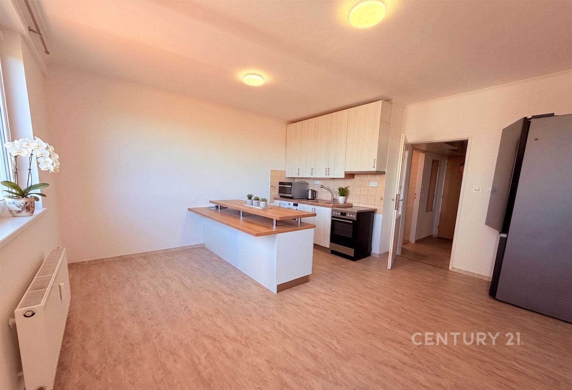 Světlá kuchyň s parketovou podlahou, vestavěné skříně a moderní spotřebiče, vedle velkého okna, nabízí pohodlí a eleganci.
