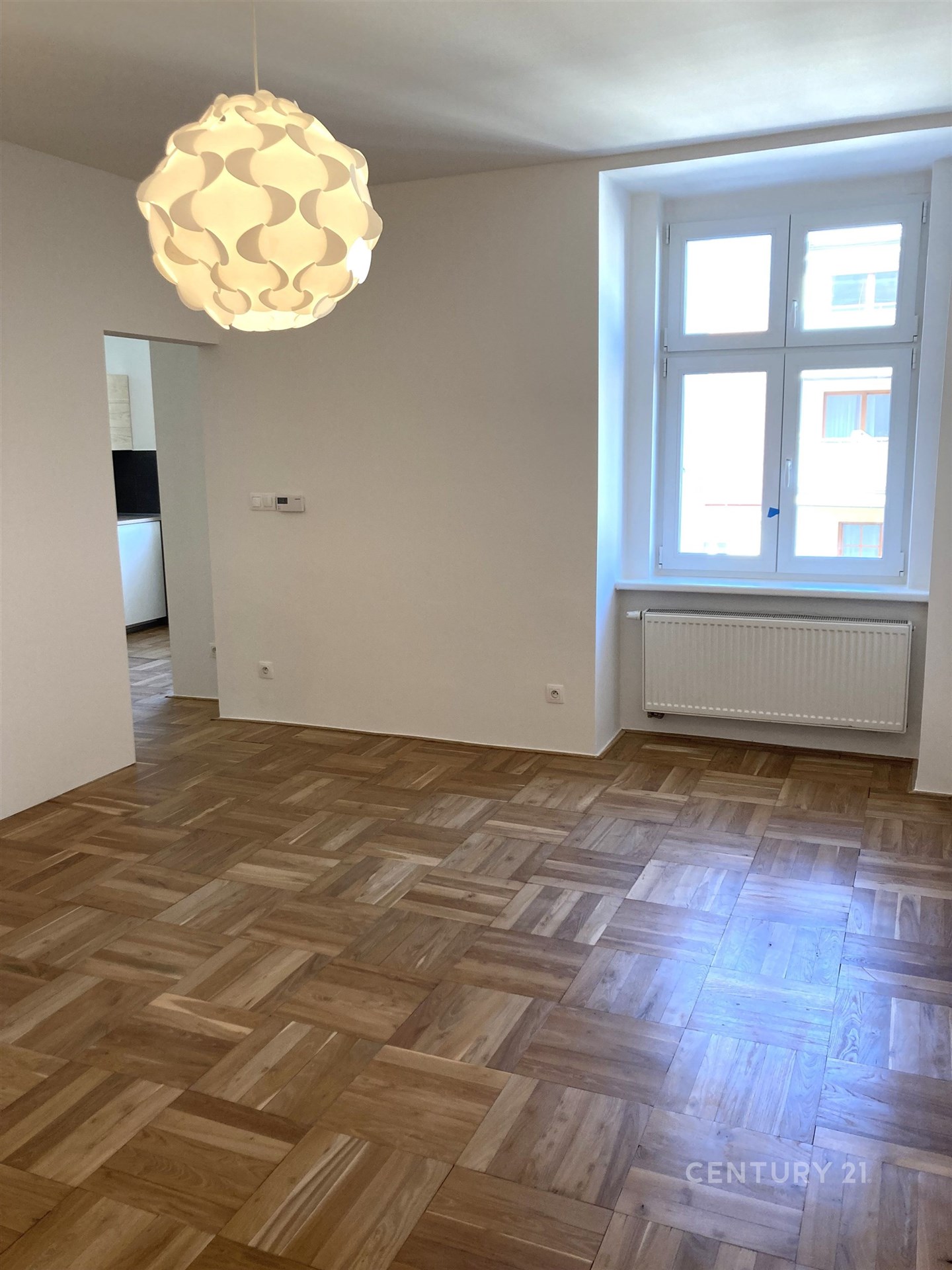 Světlý obývací pokoj s parketovou podlahou, velkými okny a moderním osvětlením, ideální pro komfortní bydlení.