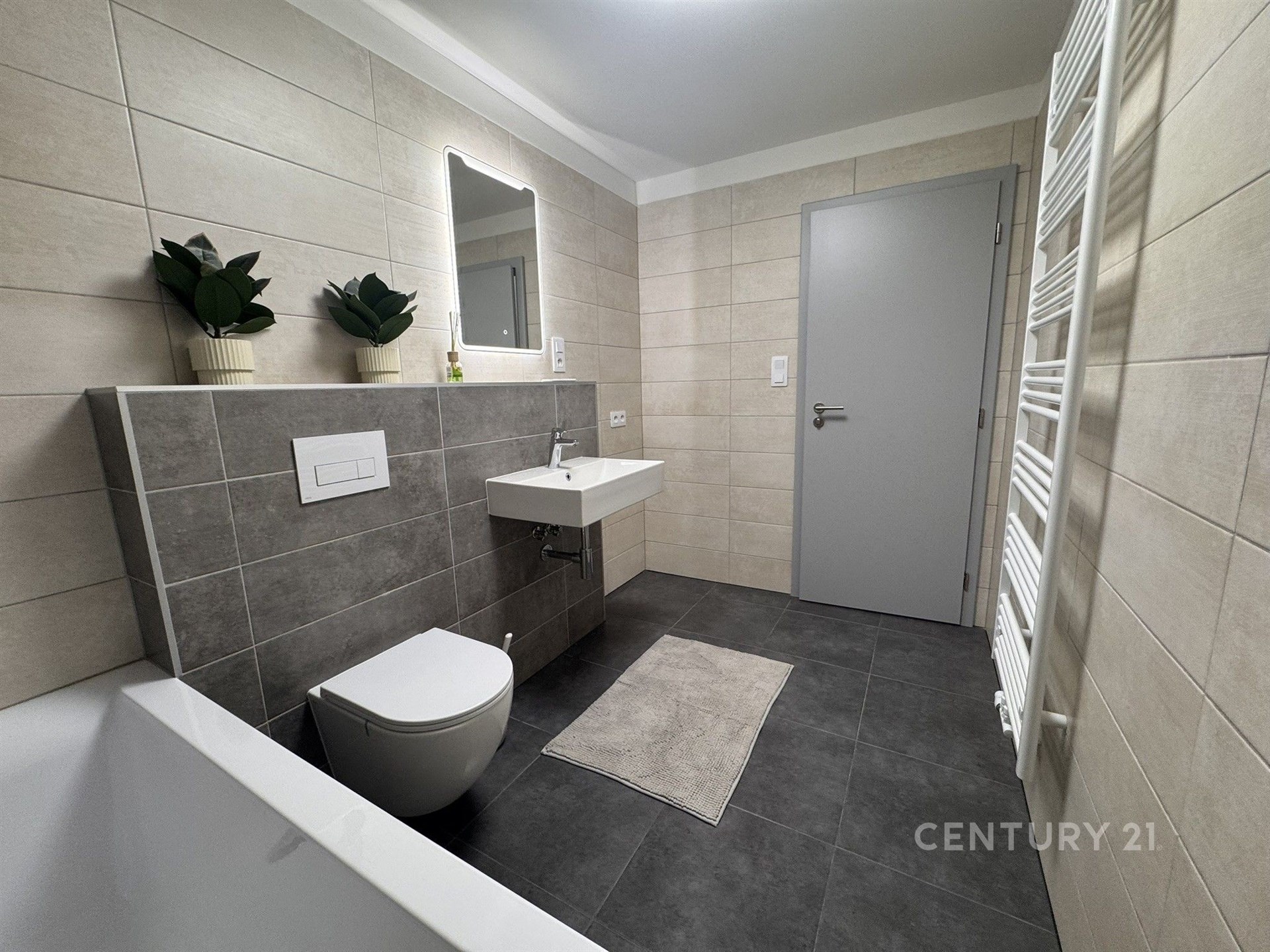 Moderní koupelna s dlažbou, světlými obklady a závěsnou toaletou, vybavená umyvadlem a velkým zrcadlem, nabízí elegantní design.