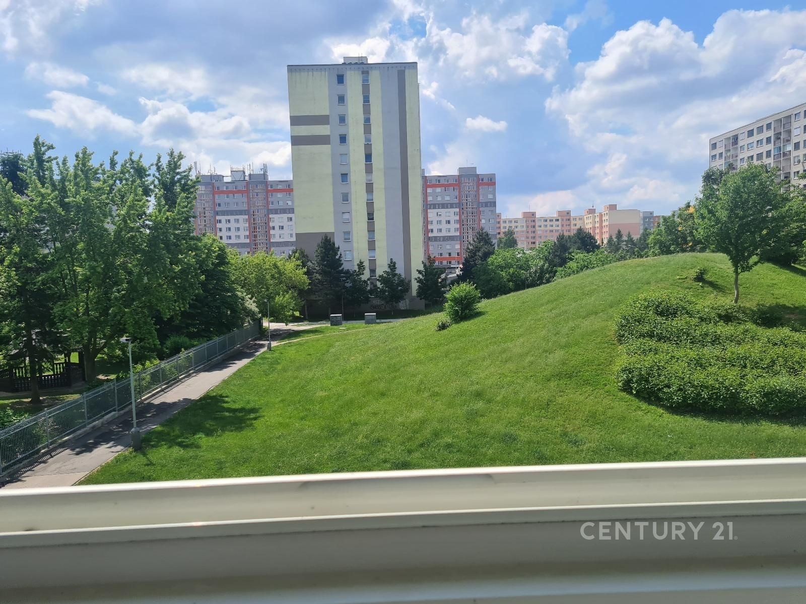 Výhled z okna na zelený park s moderními panelovými domy v pozadí, ideální pro rodiny, světlé a klidné okolí.