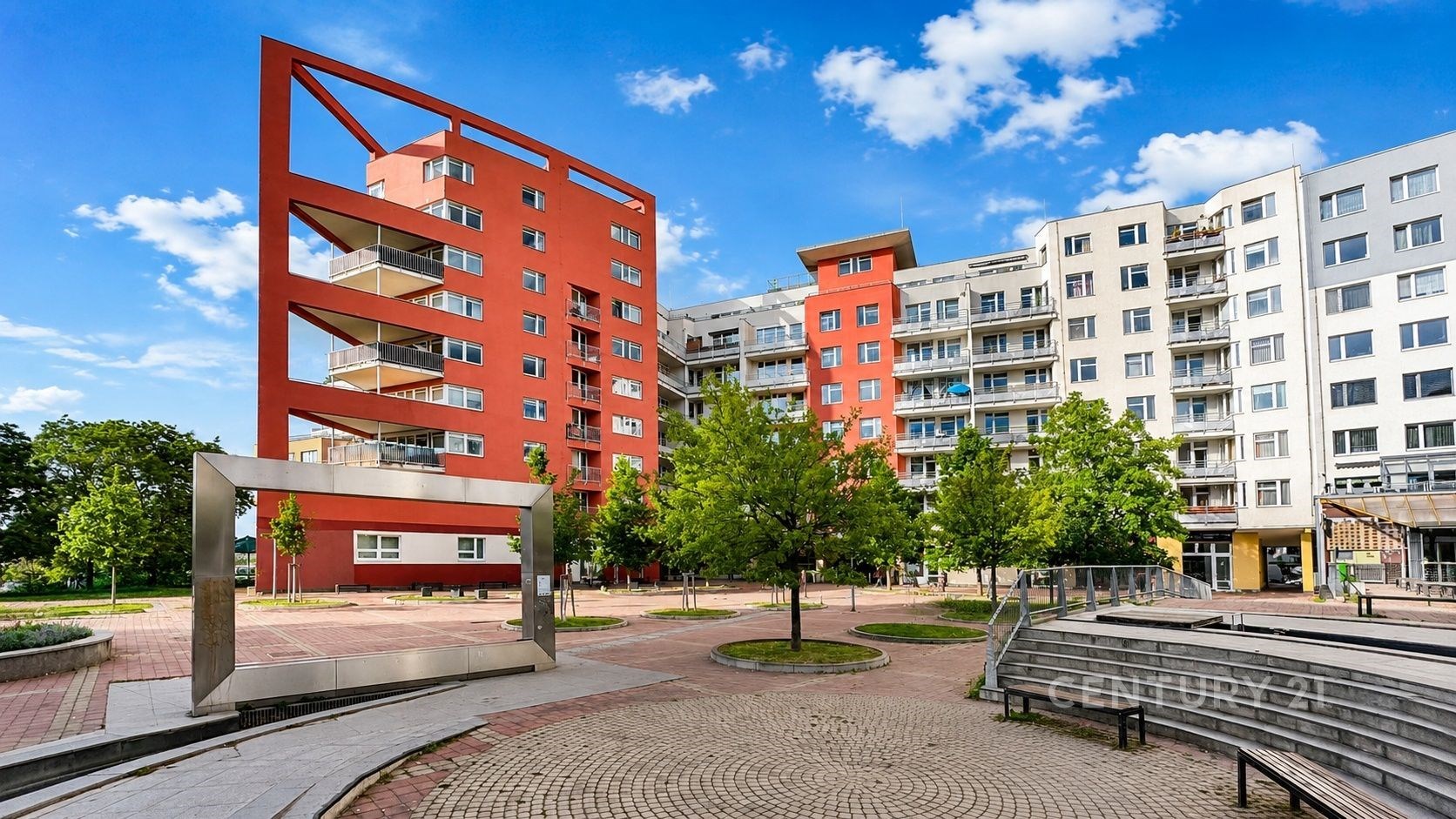 Moderní bytový komplex se světlými fasádami a balkony, obklopený zelení a dlážděnými chodníky, v rezidenční čtvrti