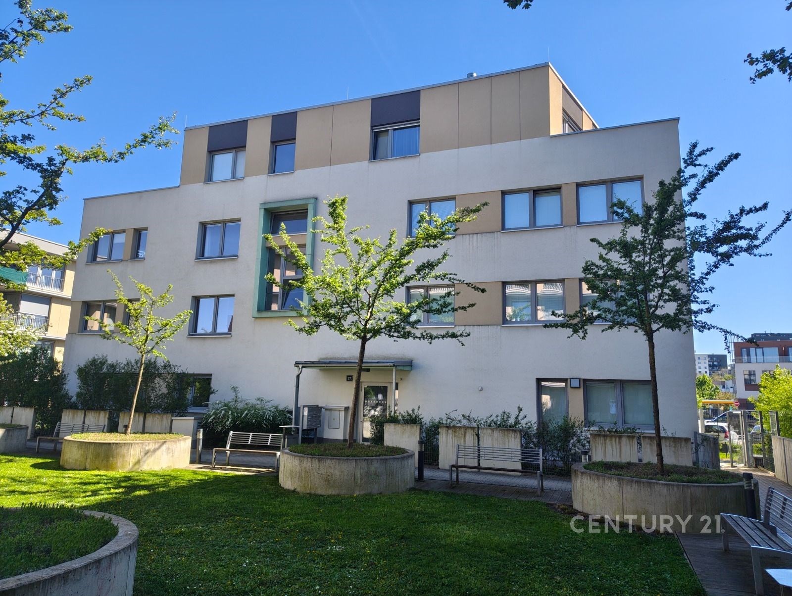 Moderní bytový dům s elegantní fasádou kombinuje světlou omítku a dřevěné obložení, obklopen upravenou zelení a klidnou městskou lokalitou.